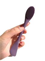 Hand holding a dual-ended silicone facial brush and spatula in purple color under natural sunlight