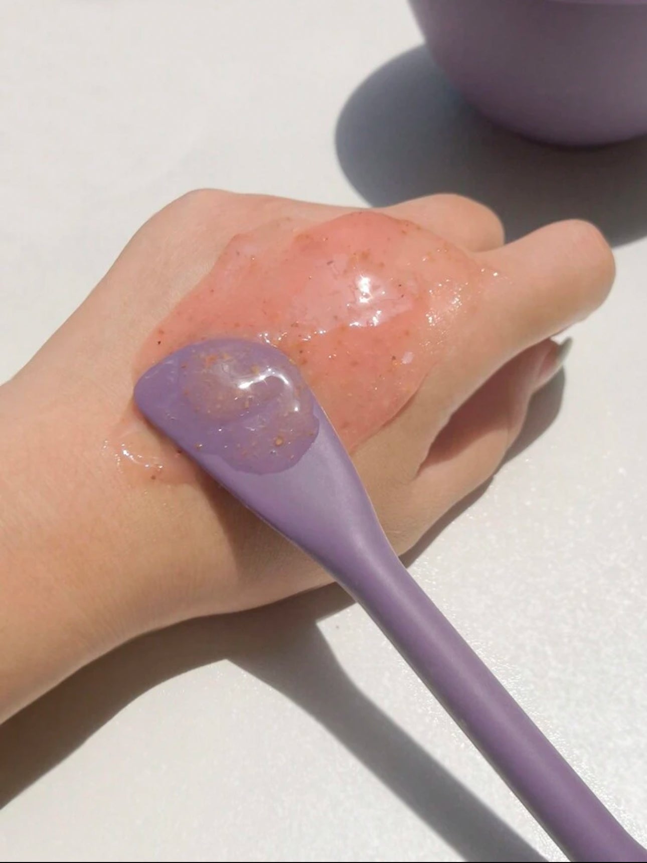 Applying gel-textured skincare product on the back of the hand using the flat side of a purple silicone spatula